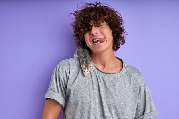 Decorative rat on joyful man's shoulders, handsome caucasian young guy doesn't afraid of exotic animals, pets. rat is crawling on t-shirt. purple wall