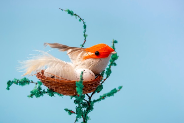 On a decorative tree a nest with eggs and a small yellow bird on a blue background close up with copy space