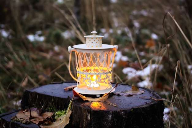 Decorative white lantern with burning candle on wet tree stump in autumn park at evening.