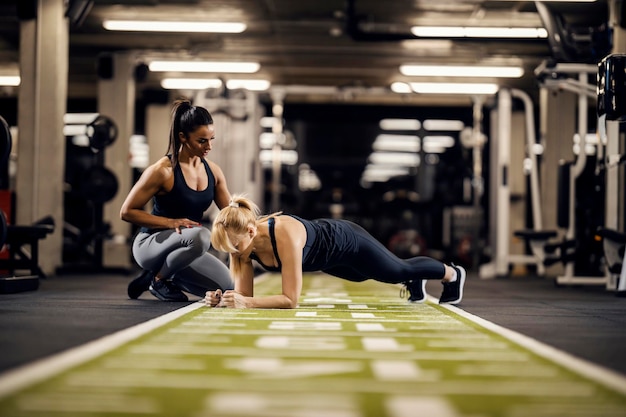 A dedicated female trainer is training sportswoman in a gym while she is ding planks