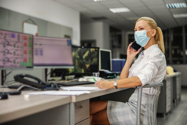 Dedicated hardworking blond female boss in suit with face mask on sitting in control room in heating plant and having important phone conversation during corona virus outbreak.