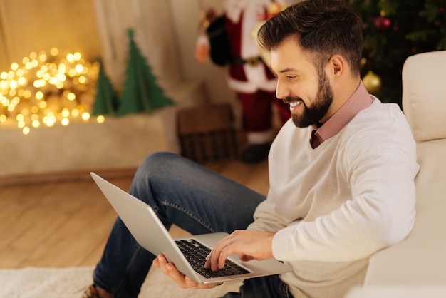 Dedicated professional. Handsome upbeat young man sitting on the floor in the room decorated for Christmas and searching for information on his laptop