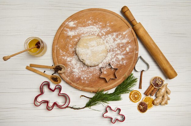 Deeg op een ronde houten plank naast een deegroller en keukengerei voor het maken van kerstkoekjes, snijmallen en gedroogde sinaasappelschijfjes en pijnboomtakken op een houten oppervlak