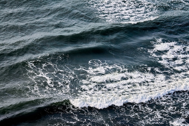 Deep blue sea waters splashing with foamy waves. Aerial view of ocean splashing waves, dark blue wavy sea waters with white foam. Water surface, sea spray. Natural background, copy space