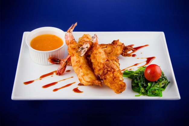 Deep-fried tiger prawns in batter served with sweet and sour sauce, nori seaweed and cherry tomatoes on a rectangular plate.