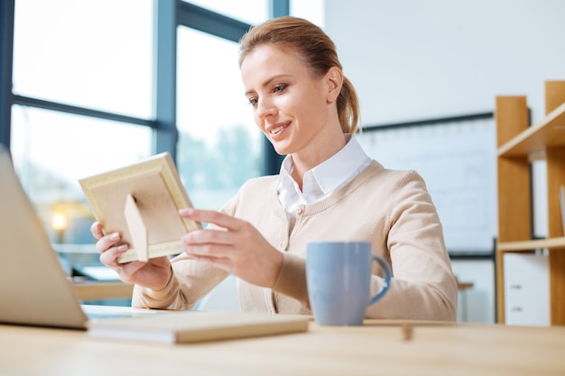 Deep in thoughts. Pleasant young woman looking at the photo in frame while holding it in both hands and expressing delight