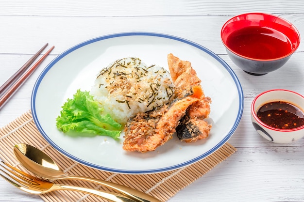 Жареный лосось подается с вареным рисом, покрытым белым кунжутом, черным кунжутом, водорослями и овощами на белой тарелке. На столе суп с соусами для макания и золотая ложка.
