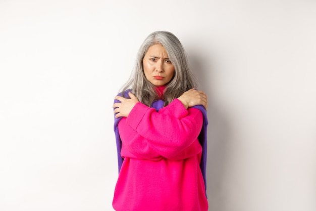 Defensive and offended asian middle-aged woman hugging body, comforting herself and looking angry at camera, frowning insulted, standing over white background.