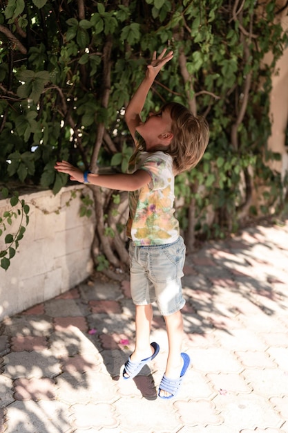 Defocused blurred photo of happy candid little kid boy of five\
years old is having fun on a summer vacation on a travel child\
jumps up trying to reach a plant on the city streets outdoor