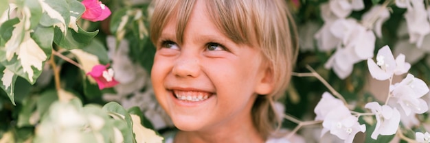 Defocused blurred portrait of face candid little happy smiling five year old blonde kid boy with green eyes in pink and white flower plants in nature children have fun summer holidays banner