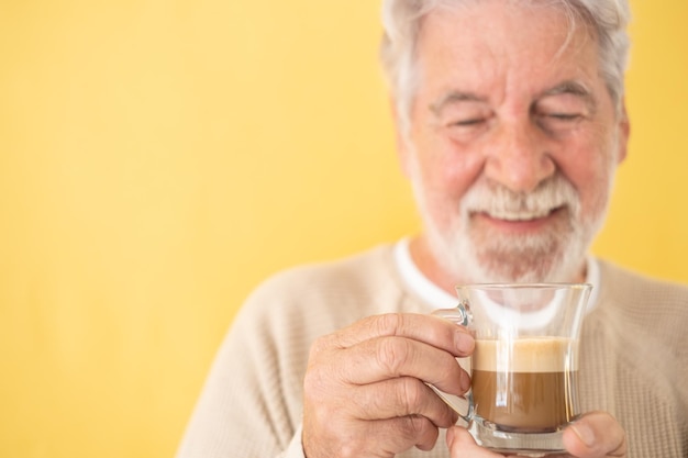 写真 黄色の背景の上に立って、コーヒーとミルク、カプチーノのガラスを保持している焦点がぼけたハンサムなひげを生やした年配の男性