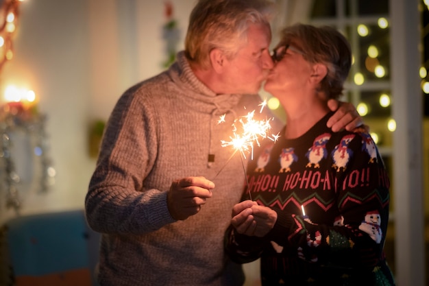 火花でクリスマスイベントを祝ってキスする焦点がぼけた年配のカップル。背景のライトとクリスマスツリー-恋にアクティブな引退した高齢者