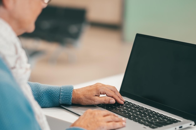 Defocused senior woman sitting at white desk using laptop typing on keyboard in business work