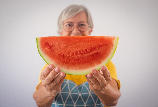 Defocused smiling caucasian old senior woman holding a slice of fresh watermelon looking at camera healthy eating lifestyle concept