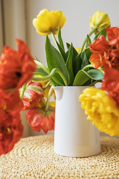 Defocused yellow red blooming tulip flowers with green stems and leaves in white ceramic pitcher on light white wall background Creative floral botany wallpaper Minimal creative greeting card