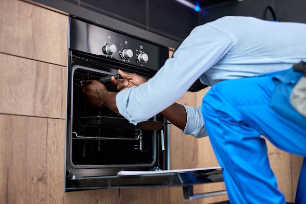 Don't delay with repair. Side view afro repairman examining oven in kitchen using tools instruments. Confident professional black repairman handyman in blue workwear overalls during work at home