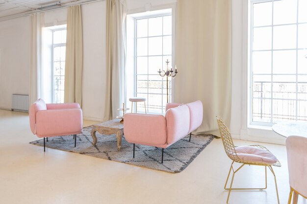 Delicate and cozy light interior of the living room with modern stylish furniture of pastel pink color and white walls with stucco moldings in daylight