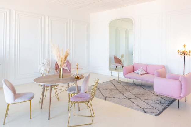 Delicate and cozy light interior of the living room with modern stylish furniture of pastel pink color and white walls with stucco moldings in daylight