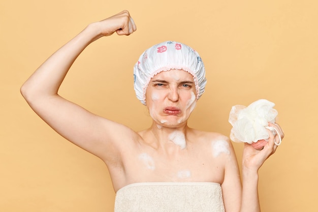 Delicate morning pampering displeased confused young woman in shower cap and wrapped in towel frowning face smelling armpit sweat isolated over beige background