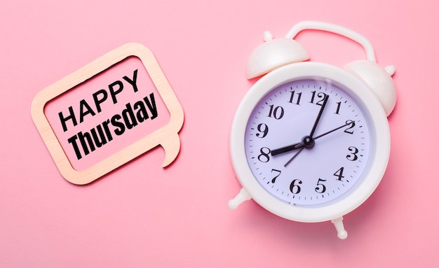 On a delicate pink background, a white alarm clock and a wooden frame with the text HAPPY THURSDAY