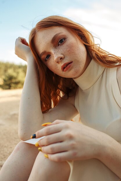 Delicate young woman with long red hair and in beige dress leaning on hand while sitting on sand