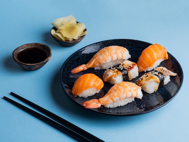Delicious appetizing sushi set served on clay plate with soy sauce and chopsticks flat lay on blue