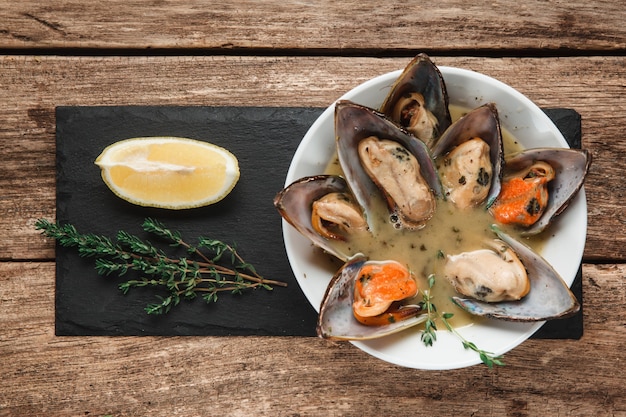 Delicious baked Mediterranian mussels served with cream sauce on black slade with herbs and lemon, top view.
