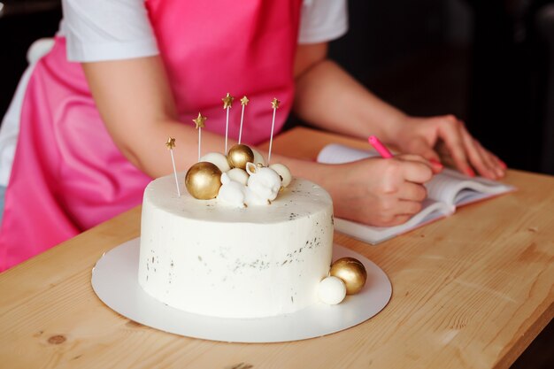 Delicious birthday cake and womans hands writing a recipe in the notebook on the table