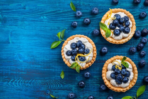 Delicious blueberry tartlets with vanilla cream on blue wooden background top view