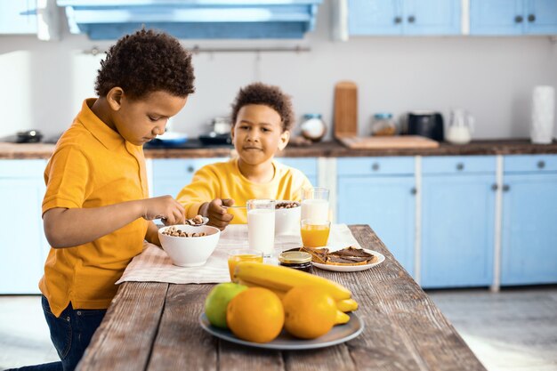 Delicious breakfast. Adorable little boy standing near the table and eating cereals for breakfast together with his elder brother