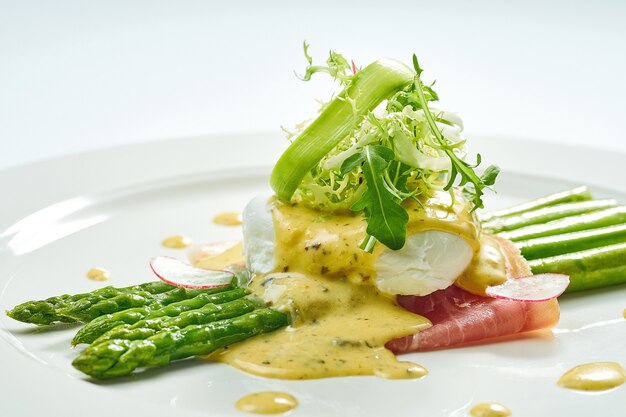 Delicious breakfast asparagus with poached egg, bacon, hollandaise sauce in a white plate. Isolated on white surface.