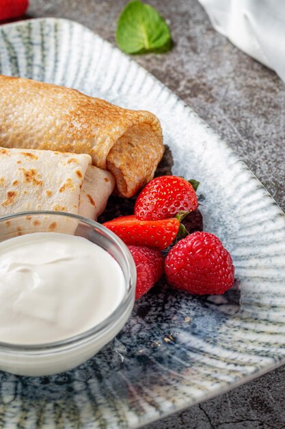 A delicious breakfast. Sweet fried pancakes filled with strawberries in a white plate with sour cream on a gray stone table with a napkin and cutlery