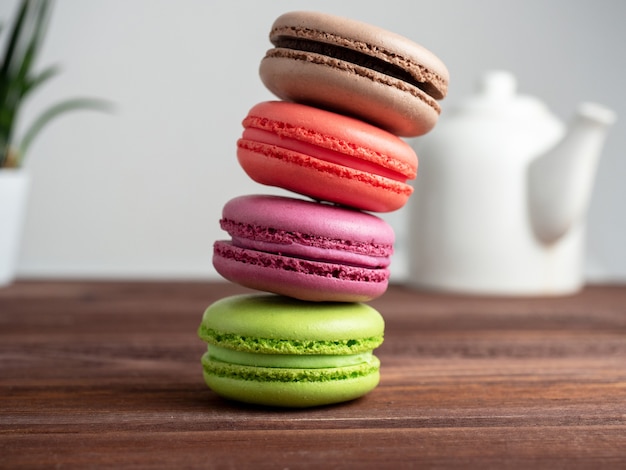 Delicious bright macaroons are stacked in a pyramid on a wooden background. In the background is a white teapot