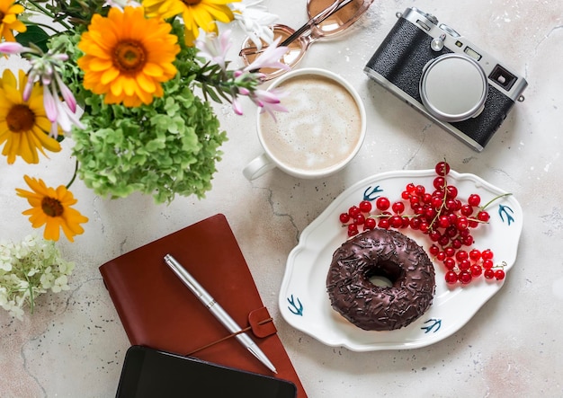 Photo delicious brunch donut with chocolate icing cappuccino bouquet of flowers camera planner on a light stone background top view