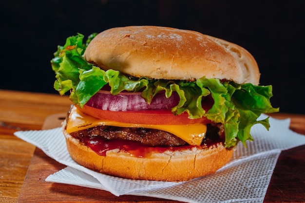 Delicious burger with lettuce, tomato and red onion and bacon on homemade bread with seeds and ketchup on a wooden surface and black background.