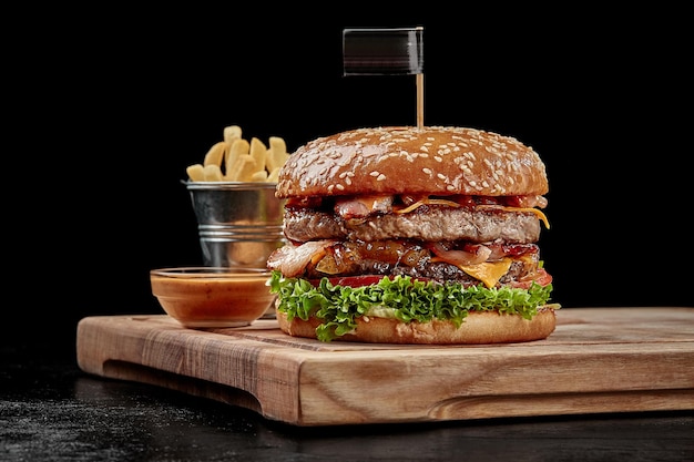 Delicious burger with two juicy pork and beef patties, fried bacon, cheese slices, caramelized onions, fresh tomatoes and greens in fluffy sesame bun served with fries and aioli sauce on wooden board