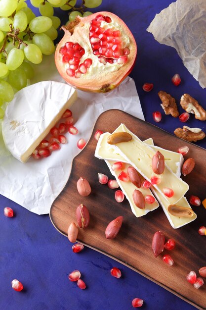 Delicious cheese on wooden board and parchment paper close up Camembert with pomegranate seeds green grapes and nuts on a blue background