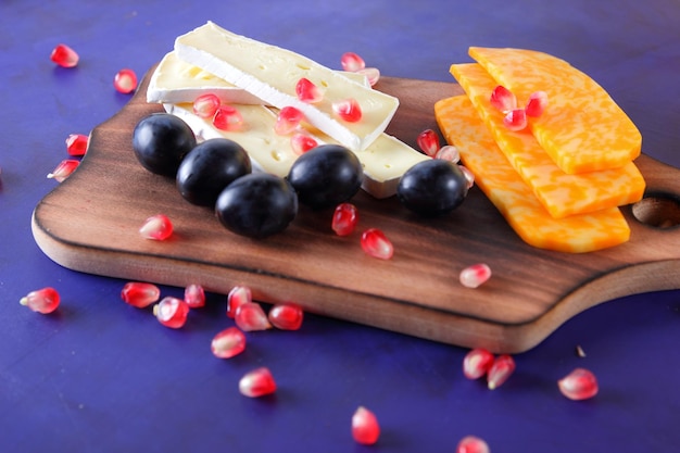 Delicious cheeses on a wooden board closeup Camembert and yellow hard cheese slices with pomegranate seeds and dark grapes on a blue background