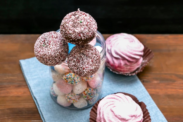 Delicious Chocolate cake pops decorated white sprinkles in jar with colorful candies sweets, pink marshmallow on napkin, wooden background with copy space, pastry, desserts on stick