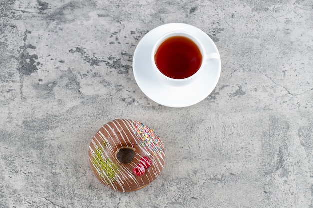 Вкусный шоколадный глазированный пончик и чашка чая на каменном фоне.