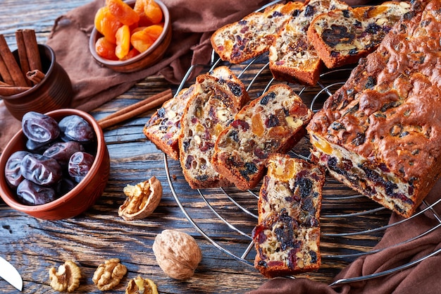 Delicious chunky dried fruits loaf cake on a wire cake stand with brown cloth, cinnamon sticks, dried apricots and date fruits on a rustic wooden table, view from above, close-up