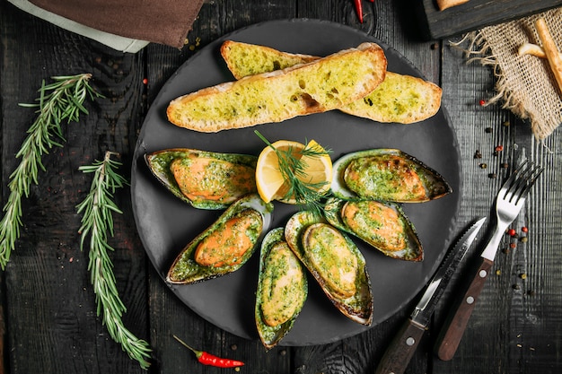 Delicious cooked mussels with sauce, lemon and garlic bread and butter on a black plate on the dark wooden background