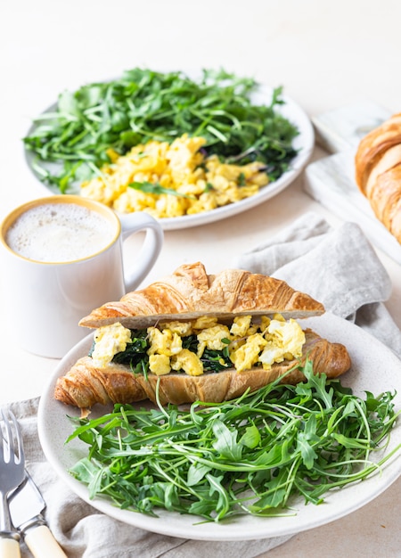 Delicious croissant sandwich with scrambled spinach eggs served with arugula on plate and coffee cup.