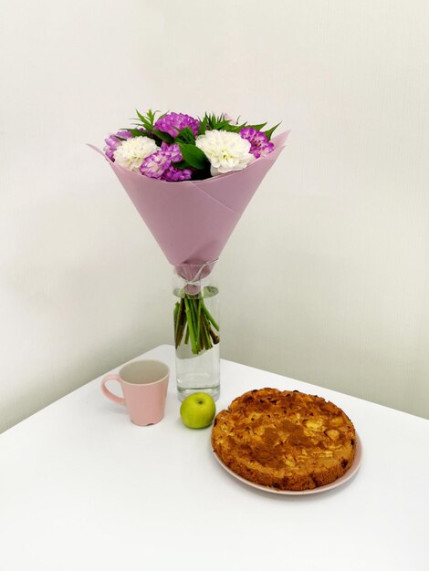 Delicious dessert apple pie on a plate, a bouquet of flowers\
and a pink mug on a white table. cozy table setting for breakfast\
and lunch. homemade baking concept.