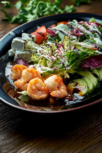 Delicious and dietary salad with grilled shrimp, feta cheese, tomatoes and avocado served in a black plate on a wooden table. Restaurant food