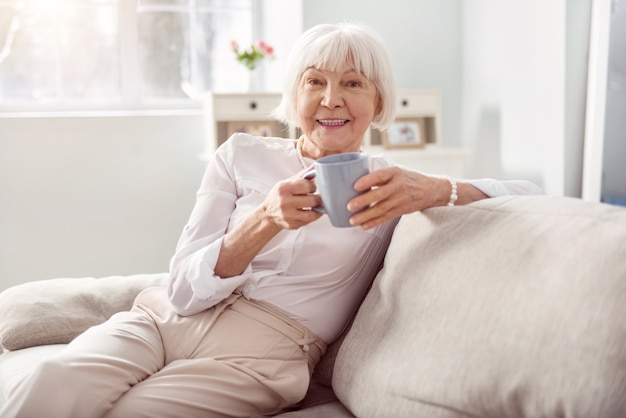 Delicious drink. Cheerful elderly woman sitting on the sofa in her living room and holding a cup of coffee while smiling