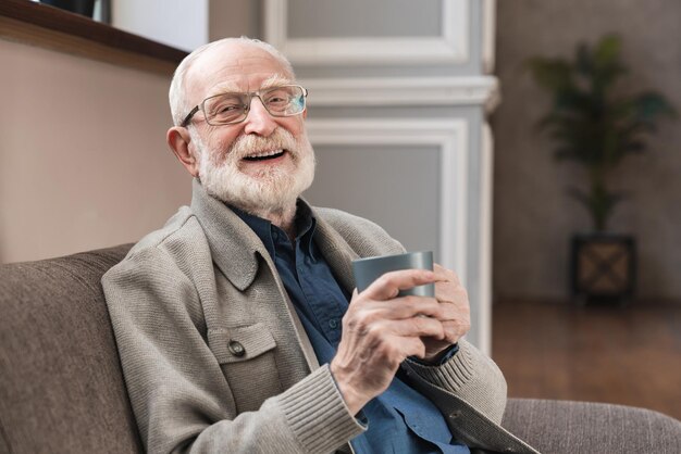 Delicious drink pleasant elderly man in spectacles sitting on\
the sofa and posing with a cup