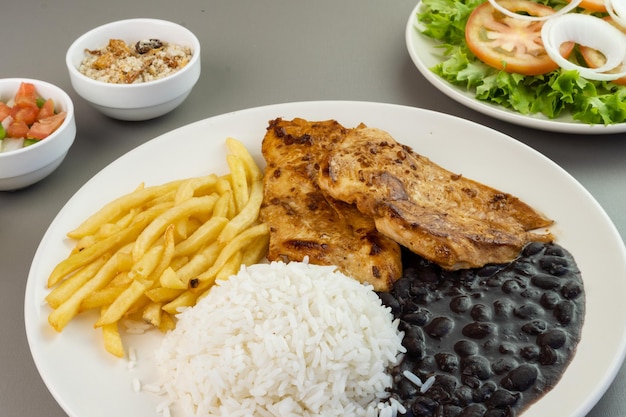 Delicious executive dish of chicken fillet rice beans french\
fries and green salad with lettuce tomato and onion accompanied by\
farofa and vinaigrette typical brazilian food selective focus