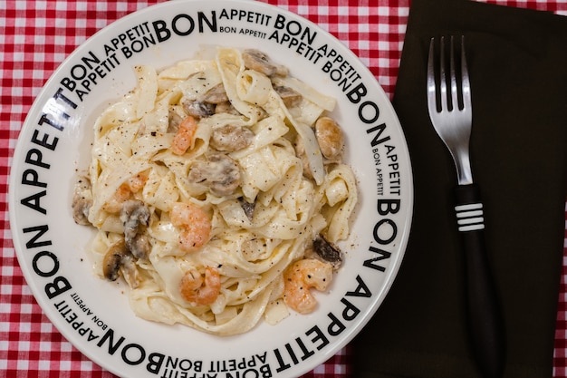 Deliziose fettuccine con salsa alla panna di funghi e gamberi su un piatto con la scritta bon appetite con tovaglia a quadretti rossa. cucina italiana.