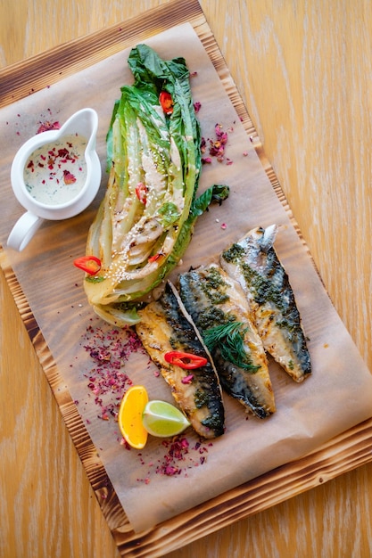 Delicious fish with spices seafood on decorated wooden board\
grilled bbq fish decorated with vegetables lemon and sauce on\
wooden table and parchment paper top view